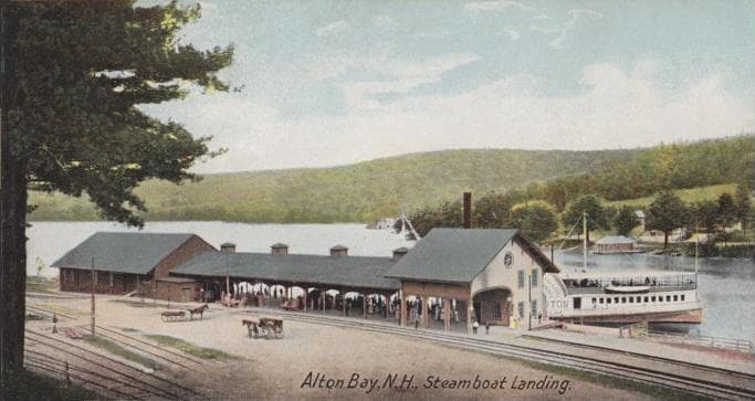A colored postcard of the Alton Bay, New Hampshire steamboat landing on Lake Winnipesaukee, showing the long railroad station building, a steamboat at the dock, and tracks running along the lakefront.