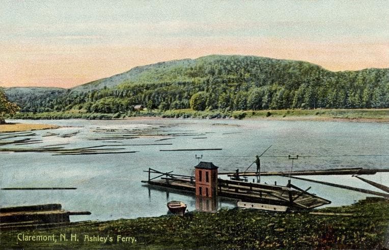 A colored postcard of Ashley's Ferry on the Connecticut River at Claremont, New Hampshire, showing a small wooden ferry platform with a man standing on it handling a long pole, logs floating in the river, and forested hills on the Vermont side opposite.