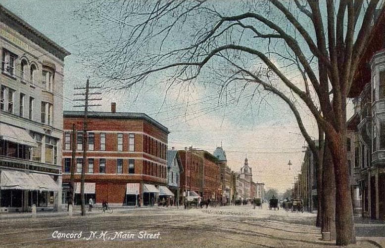 A colored postcard of Main Street in Concord, New Hampshire, with brick commercial buildings on either side, a wide bare elm tree in the foreground reaching across the frame, the State House dome visible in the distance, and small horse carts in the street.