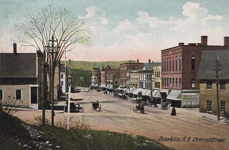A colored postcard of Central Street in Franklin, New Hampshire, ca. 1910, showing brick commercial buildings on either side of a wide dirt street with awnings, telephone poles, a horse cart, and a single bare tree in the foreground.