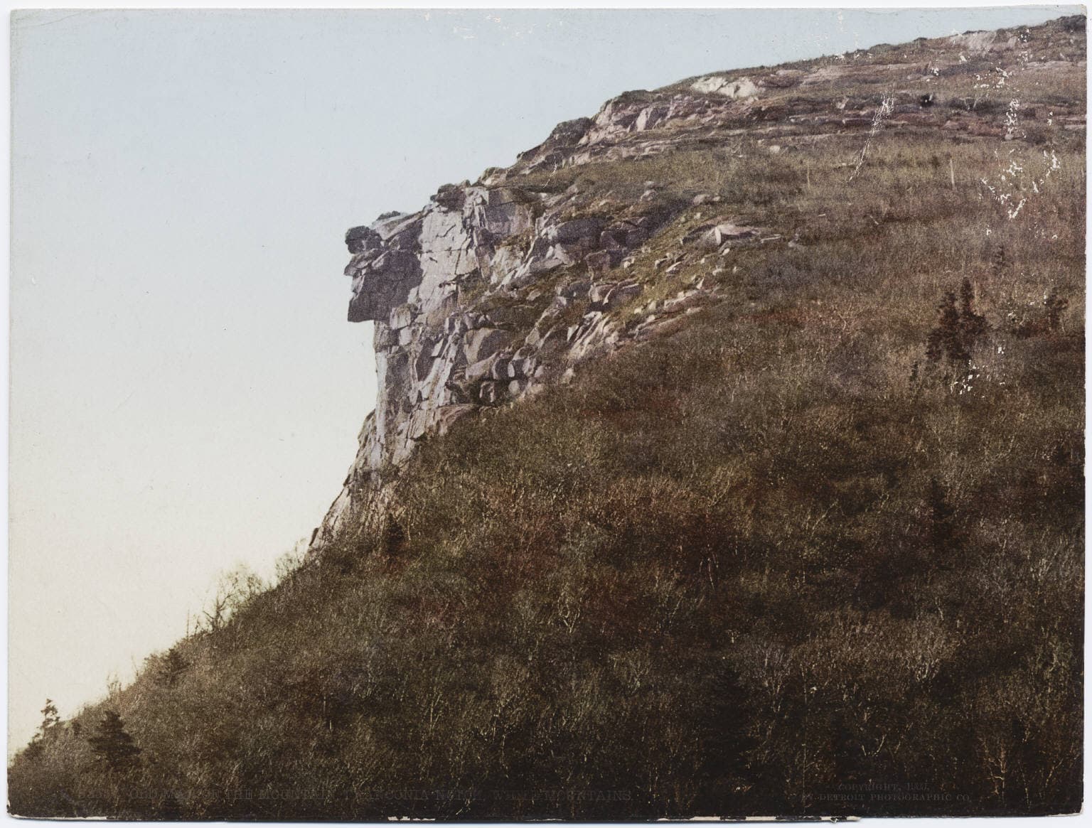 A hand-tinted Detroit Publishing photochrom, ca. 1900, of the Old Man of the Mountain rock formation in Franconia Notch, New Hampshire, showing the granite profile in cliff against an open sky with green hillside below.